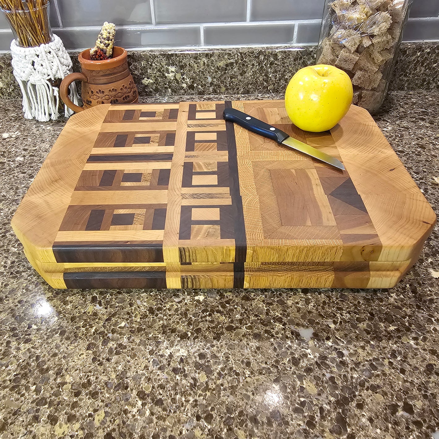 A wooden cutting board with a geometric pattern is placed on a speckled granite countertop. On the cutting board, there is a yellow apple and a small knife with a black handle. In the background, there is a ceramic mug with a handle, a decorative item with dried flowers, and a glass container filled with brown sugar cubes. The cutting board features a mix of light and dark wood tones arranged in a visually appealing design